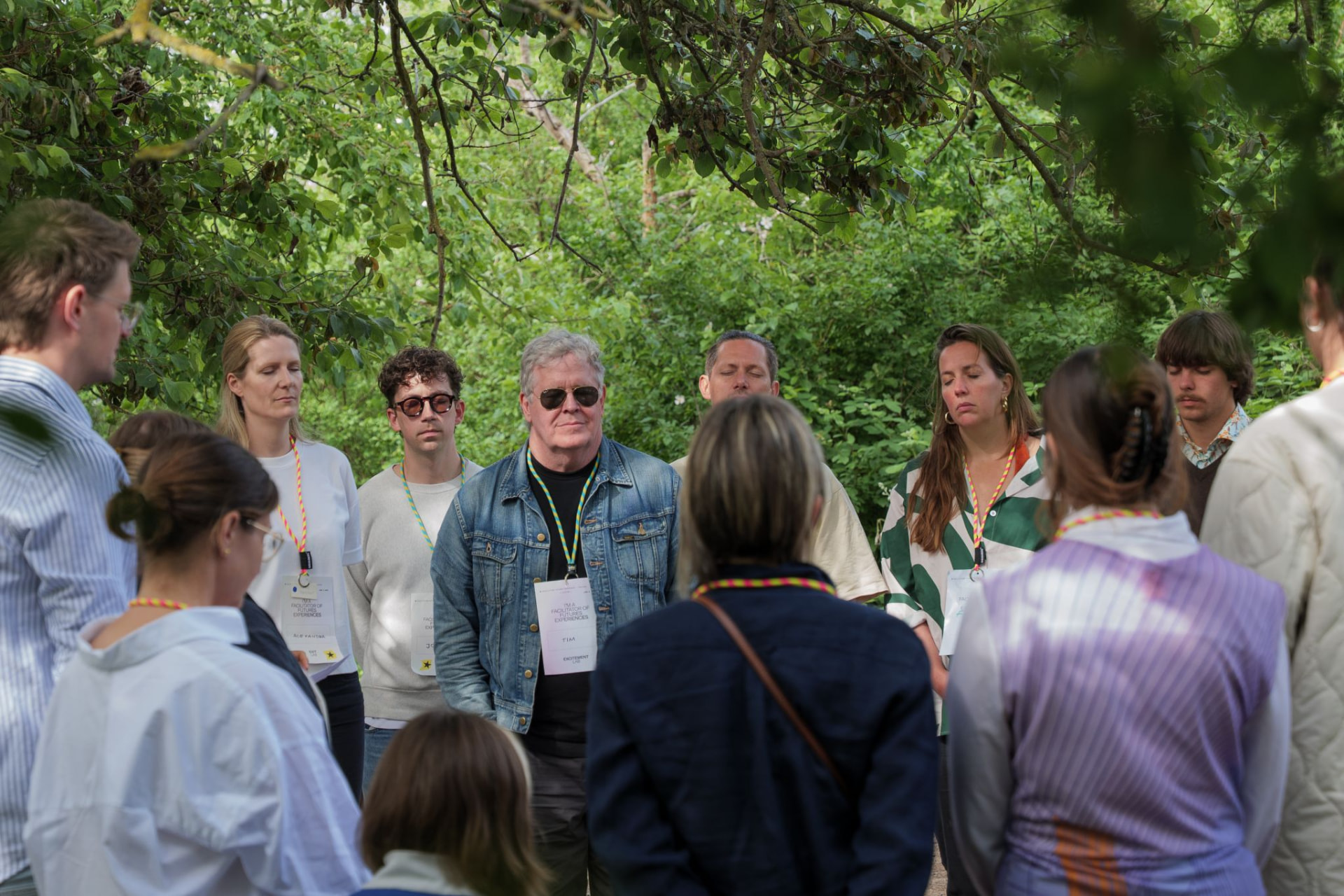 A circle of participants standing together under green trees during a gathering
